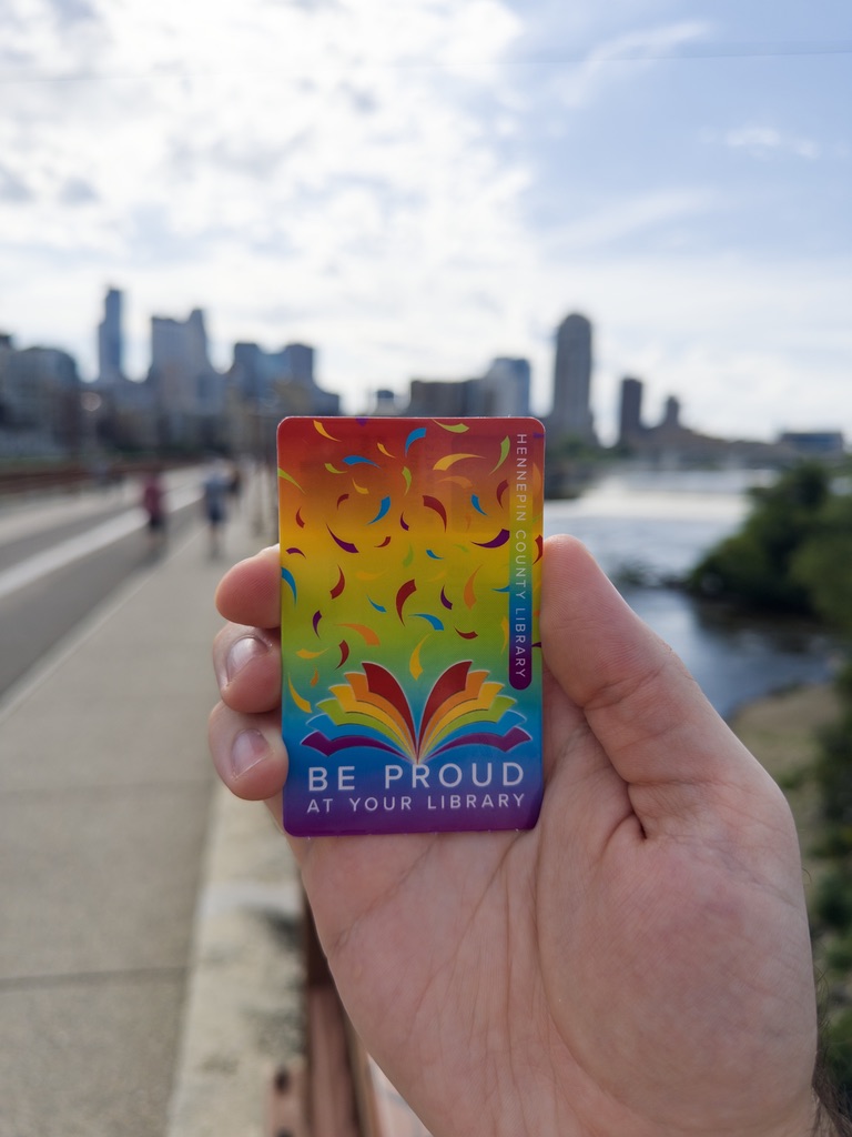 Holding my Hennepin County Library card out in front of me, with the skyline of downtown Minneapolis in the background. The library card has a rainbow design, with the text "Be proud at your library"