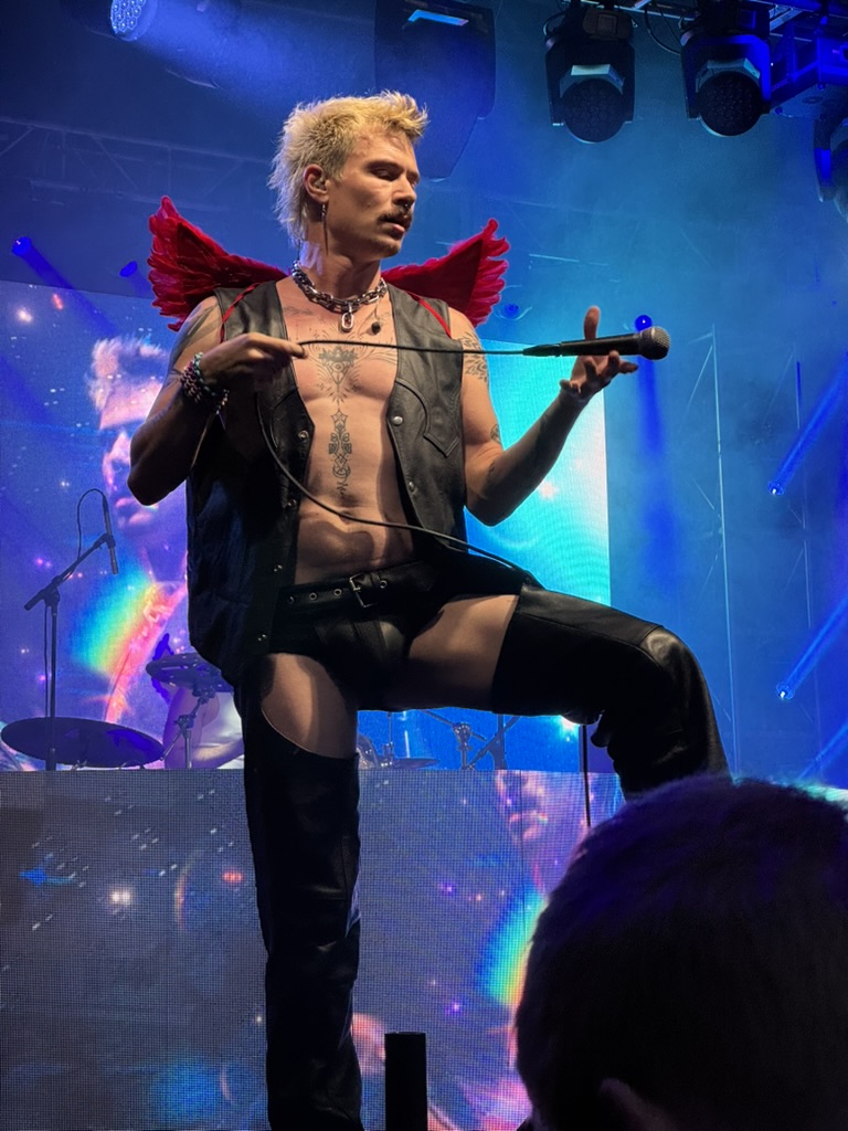 Zee Machine performing on stage wearing a leather vest, shirtless, and red wings