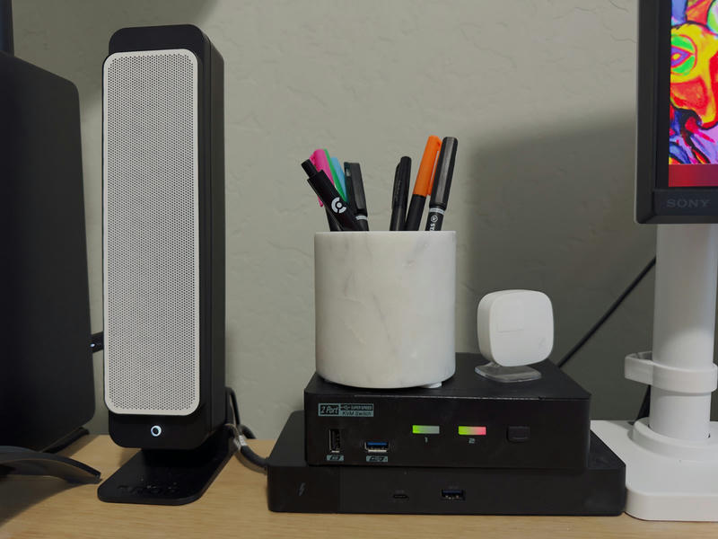 To the left is the Drop BMR1 speaker, which is slender with a white mesh grille. To the right of the speaker is the KVM, sitting on top of a Thunderbolt 3 dock. On top of the KVM is a white marble pen holder, with various pens, one of which displays the Clerk logo.
