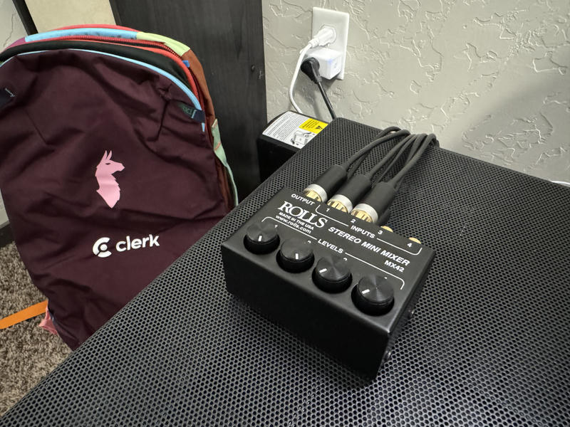 The ROLLS MX42 Stereo Mini Mixer sitting on top of a black mesh grille. To the left is a burgandy backpack with the Clerk wordmark on it.